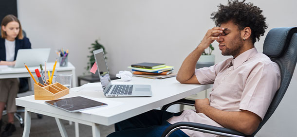 procrastinação no trabalho, homem negro com a mão no rosto de olhos fechados no escritorio, ao fundo uma mulher focada trabalhando