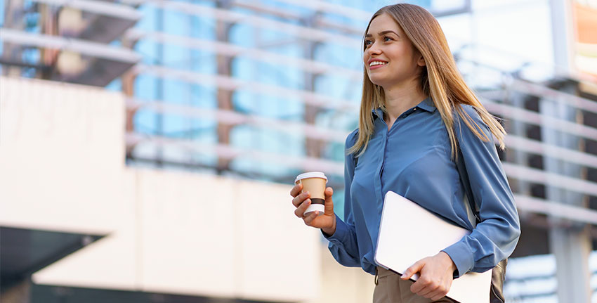 carreira em y, jovem loira sorrindo segurando um laptop e café, vestindo uma camisa azul suave sobre um edifício moderno