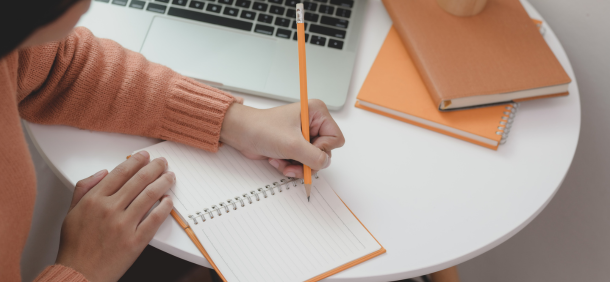 uma pessoa anota em um caderno diante de um notebook e materiais organizados sobre uma mesa branca