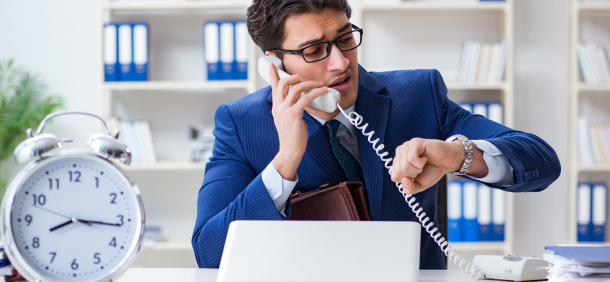 Administração do tempo homem de negócios aparenta estresse ao falar ao telefone e checar o relógio simultaneamente, cercado por objetos de trabalho e um grande despertador