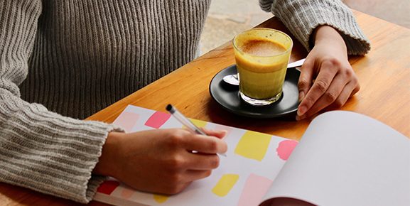 mãos segurando caneta, fazendo sua organização pessoal em caderno, xicara de café a mesa